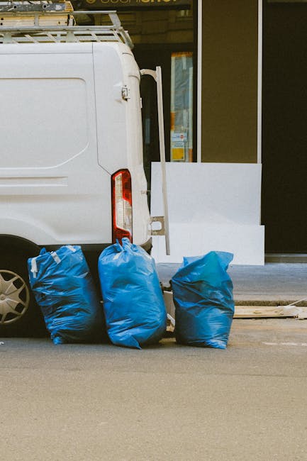 The image shows the rear side of a white commercial van, positioned near the curb on an urban street. The van's back door is partially visible, with a vertical handle and some hinge details, and it appears to be parked close to a building entrance with large glass windows and doors. In the foreground, three large blue plastic rubbish bags are neatly arranged on the pavement, filled with waste materials, likely part of a private rubbish collection service provided by companies like rubbishclearanceelephantandcastle.co.uk. The bags are slightly crinkled, with some peaks and folds, and are situated on a smooth asphalt road surface. To the right, a section of the sidewalk can be seen, with a white concrete step leading to the building entrance. The setting suggests on-site clearance or alternative waste handling methods rather than local authority collection, emphasizing the context of commercial or private rubbish removal services focused on quick, independent rubbish pickup near Elephant Park. The scene is illuminated evenly, with natural light indicating a daytime setting, and the overall environment maintains a neutral aesthetic suitable for waste management documentation or service descriptions.