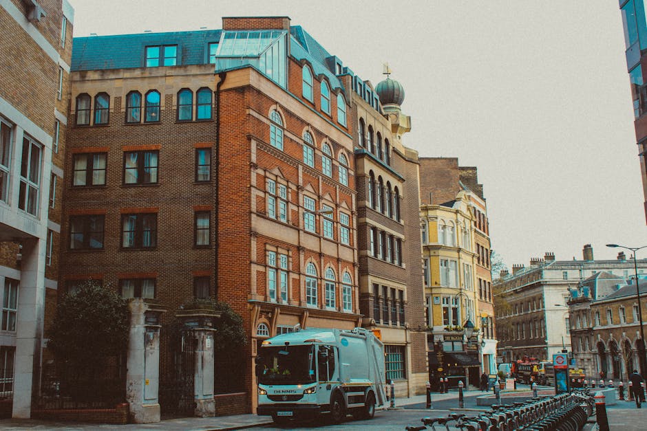A street scene in an urban area shows a row of historic multi-storey buildings with brick and ornate facades, some with decorative arches and large windows, beside modern structures. A small white waste collection truck is parked on the pavement near a gated entrance with stone pillars, likely part of a private waste management service. The truck is positioned to the left of the image, close to a black iron fence and some greenery. To the right, several bicycle racks are lined up along the sidewalk, and a few bollards prevent vehicle access to the pedestrian area. Above, the sky is overcast, casting diffused light over the scene, with some reflections visible in the building windows. Overall, the environment suggests a busy city street where independent rubbish collection or on-site clearance may be utilized to manage waste, aligning with private disposal options outside local authority services. The scene captures the typical street-level view of a central London neighborhood with a mix of historic architecture and modern urban elements.
