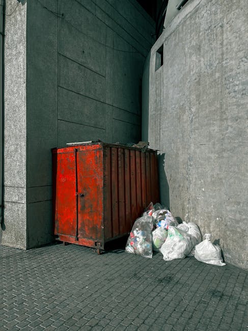 A street scene in an urban area shows a row of historic multi-storey buildings with brick and ornate facades, some with decorative arches and large windows, beside modern structures. A small white waste collection truck is parked on the pavement near a gated entrance with stone pillars, likely part of a private waste management service. The truck is positioned to the left of the image, close to a black iron fence and some greenery. To the right, several bicycle racks are lined up along the sidewalk, and a few bollards prevent vehicle access to the pedestrian area. Above, the sky is overcast, casting diffused light over the scene, with some reflections visible in the building windows. Overall, the environment suggests a busy city street where independent rubbish collection or on-site clearance may be utilized to manage waste, aligning with private disposal options outside local authority services. The scene captures the typical street-level view of a central London neighborhood with a mix of historic architecture and modern urban elements.