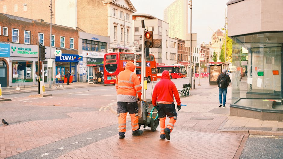 Two waste collection workers in bright orange high-visibility uniforms, with reflective stripes, are walking together on a city sidewalk in Elephant and Castle. One worker, wearing a hooded jacket, is pushing a wheeled bin, while the other, also dressed in a hooded jacket, walks alongside. Behind them, there are several red double-decker buses parked near a traffic light showing a red signal. The background includes a variety of commercial buildings, such as a building with a sign for Yorkshire Building Society, and storefronts with glass windows. One pedestrian is seen walking away in the distance, and a bench is positioned further along the sidewalk. The scene is captured in natural daylight with clear weather, reflecting a typical urban environment where private waste collection may be part of on-site clearing or alternative rubbish management, external to local authority services. Overall, the image emphasizes street-level activity related to waste disposal and urban maintenance in a busy commercial area.