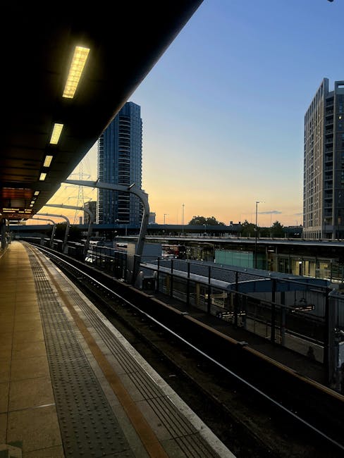 View of an urban railway station platform at sunset, with a concrete surface featuring tactile paving strips near the edge for safety. Above, a dark overhanging structure with evenly spaced rectangular lighting panels illuminates part of the platform. In the background, there are modern high-rise buildings with glass and metal facades, indicating a city centre setting. An elevated railway track runs parallel to the platform, supported by metal railings and columns, while a multi-storey building with large windows is visible on the right. The scene captures a quiet moment in a metropolitan area, with a warm, fading light creating a contrast between the illuminated platform and the cool tones of the surrounding buildings. The setting suggests a modern urban environment where urban infrastructure and development are prominent, aligning with the context of city-based public transport and private waste disposal operations managed by local services such as rubbish clearance companies including rubbishclearanceelephantandcastle.co.uk.