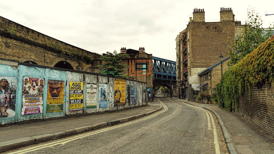 A narrow urban street scene featuring a gently curved paved road with double yellow lines along the edge, bordered by a tall brick wall covered in multicoloured posters on the left side and a neatly maintained hedge on the right side. In the background, a blue metal bridge spans across the street, and several brick buildings with chimneys line both sides of the road. The sky is overcast with grey clouds, casting diffuse natural light over the scene. The street appears to be in a quiet area, with no vehicles or pedestrians visible. This setting may relate to private waste or rubbish collection services like those offered by rubbishclearanceelephantandcastle.co.uk, especially given the urban context and proximity to structures that might generate waste needing clearance. The overall environment has a slightly aged, industrial feel, with textures from brick, metal, and weathered posters contributing to a sense of a typical city street often serviced for rubbish removal or clearance activities.