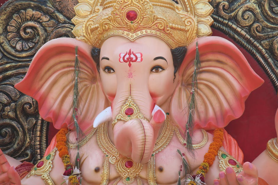 A detailed close-up of a large, intricately decorated statue of the Hindu deity Ganesha, featuring a pinkish-orange elephant head with prominent ears, a curved trunk, and expressive eyes. The statue is adorned with gold embellishments, including a decorative headpiece with a red gemstone at the center, and layered gold jewelry around the neck. The surface of the statue displays a smooth, polished finish, with fine detailing on the facial features and ornaments. The background consists of ornate, dark-colored wood carvings with scrolling patterns, providing a rich contrast to the vibrant colors of the statue. Small floral garlands made of bright orange marigolds drape over the shoulders, and strips of fabric hang from the ears, adding to the ceremonial appearance. The overall lighting highlights the shiny, reflective surfaces and textures, emphasizing the craftsmanship and vibrant spirituality. This imagery reflects traditional religious iconography commonly seen during celebrations, illustrating the cultural significance of such statues, which are often prepared for processions or rituals related to Hindu practices and festivals where independent collection or private disposal services, such as those offered by rubbish clearance specialists, may be employed for respectful handling and disposal of damaged or used statues afterward.