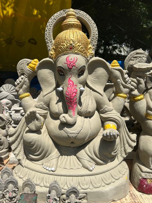 A detailed stone statue of the Hindu deity Ganesha, prominently featuring an elephant head with large ears and a curved trunk painted pink. The statue depicts Ganesha seated on a decorative lotus base, with multiple arms holding various symbolic objects. The idol is adorned with a golden crown, a red tilak on the forehead, and yellow accents on the arms and headgear. The surface of the statue has a smooth, polished finish with intricate carvings and textures that highlight the craftsmanship. In the background, other smaller statues and figures are visible, along with some foliage and shadows cast by trees, suggesting that the statue is situated outdoors. The lighting emphasizes the contrast between the light-colored stone and the darker surroundings, creating a vibrant and detailed visual impression typical of religious or cultural sculptures often seen in outdoor shrines or temple areas. This visual detail aligns with themes of spiritual iconography and traditional craftsmanship often associated with religious statues, yet in the context of rubbish removal services, it highlights the importance of respectful handling and care when managing outdoor cultural artifacts.