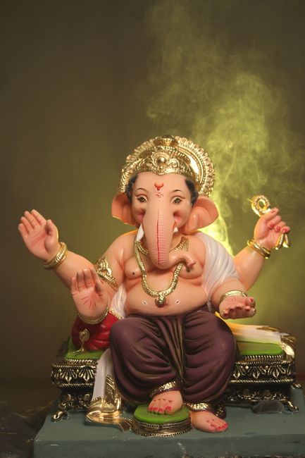 A detailed close-up of a large, intricately decorated statue of the Hindu deity Ganesha, featuring a pinkish-orange elephant head with prominent ears, a curved trunk, and expressive eyes. The statue is adorned with gold embellishments, including a decorative headpiece with a red gemstone at the center, and layered gold jewelry around the neck. The surface of the statue displays a smooth, polished finish, with fine detailing on the facial features and ornaments. The background consists of ornate, dark-colored wood carvings with scrolling patterns, providing a rich contrast to the vibrant colors of the statue. Small floral garlands made of bright orange marigolds drape over the shoulders, and strips of fabric hang from the ears, adding to the ceremonial appearance. The overall lighting highlights the shiny, reflective surfaces and textures, emphasizing the craftsmanship and vibrant spirituality. This imagery reflects traditional religious iconography commonly seen during celebrations, illustrating the cultural significance of such statues, which are often prepared for processions or rituals related to Hindu practices and festivals where independent collection or private disposal services, such as those offered by rubbish clearance specialists, may be employed for respectful handling and disposal of damaged or used statues afterward.
