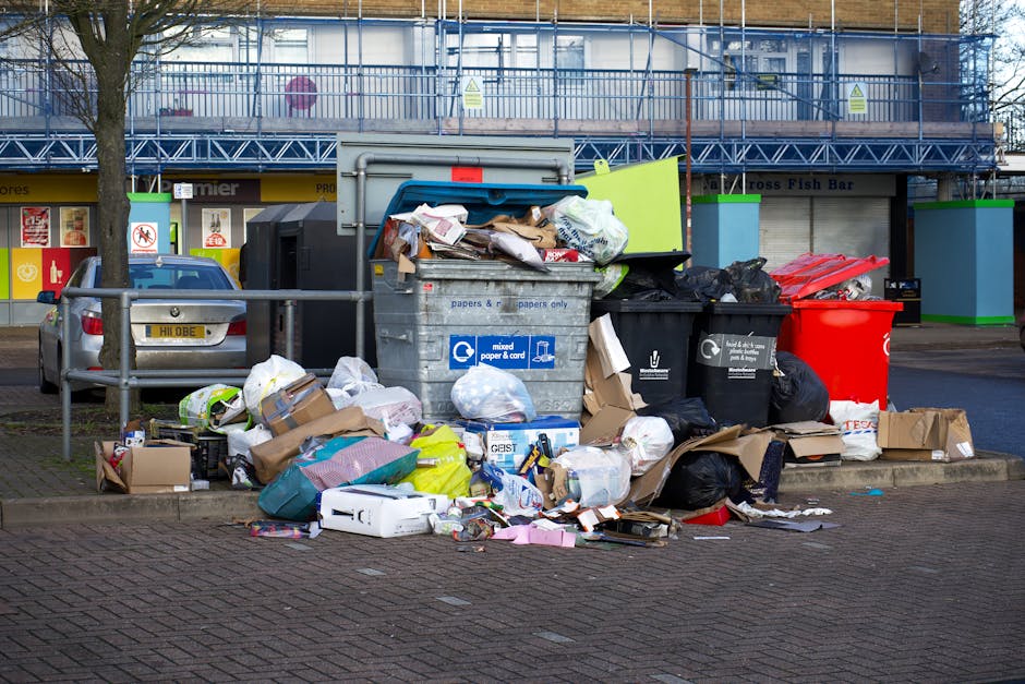 A large accumulation of mixed waste and rubbish bags is scattered around a metal recycling bin in a parking lot, with some bags and cardboard boxes on the paved surface in front. The recycling bin, labeled for mixed paper and card, is overflowing with crumpled paper, cardboard, and plastic wrapped items. Surrounding the bin are various black trash bags, a bright red waste container, and additional cardboard boxes, some partially crushed or torn. In the background, part of a parked silver car with a registration plate, along with a multicoloured building featuring shop fronts, is visible. A metal railing separates the rubbish area from the parked vehicle, and a leafless tree is positioned to the left of the scene. The environment appears to be an urban or commercial area with a grey overcast sky, and the scene reflects a private rubbish collection point, potentially managed by independent waste removal services like those offered by rubbishclearanceelephantandcastle.co.uk, suitable for on-site clearance or alternative waste handling outside municipal collection.