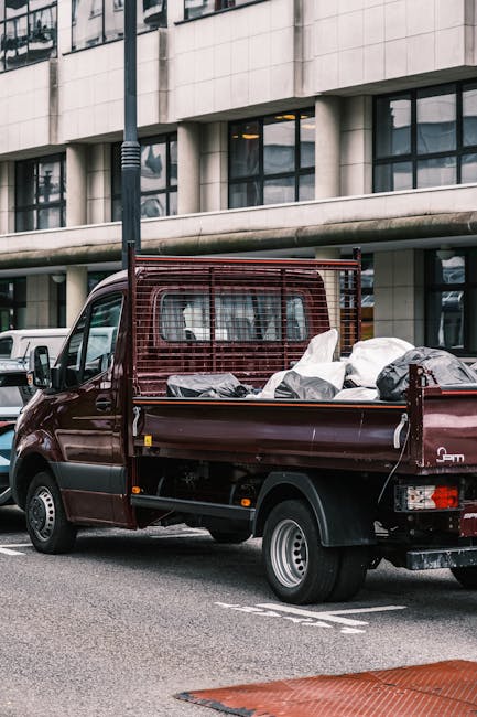 A dark red flatbed pickup truck parked on a city street in front of a modern, multi-storey office building with large glass windows and concrete facade. The truck's bed is loaded with various types of refuse, including black and white plastic bags, cardboard boxes, and other debris. The cargo appears to be loosely stacked, with some bags slightly bulging and materials spilling over the sides. A metal mesh barrier is positioned behind the cab, providing structural support and protection. The vehicle is part of a professional waste removal service, likely engaged in private rubbish clearance, as it is situated in an urban environment with nearby parked cars and quiet street surroundings. The weather appears overcast, casting diffuse light over the scene, emphasizing the textures of the scattered rubbish, the truck's smooth painted surface, and the reflective glass windows of the building in the background. This setup exemplifies typical on-site waste collection undertaken by independent rubbish clearance companies in city centres, aligning with non-local authority disposal methods.