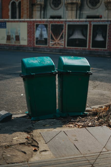 Two green plastic wheelie bins with hinged lids featuring white text are positioned side by side on a concrete sidewalk near the edge of the road, with small scattered leaves and dirt around their bases. The bins are placed on a drainage grate, and behind them is a paved street with a slight incline. In the background, there is a brick wall with large window displays that appear to be covered or blurred, and a beige building with decorative architectural features. The scene is illuminated by natural daylight, creating mild shadows cast by the bins and nearby objects. This setting suggests an area where private waste collection may occur, consistent with independent rubbish removal services, and reflects typical street-level waste management practices often encountered outside of municipal collection schemes. The presence of a company, such as Rubbish Clearance Elephant and Castle, may be implied, though no signage or branding is visible on the bins or surroundings, aligning with the context of alternative waste disposal options in urban environments.
